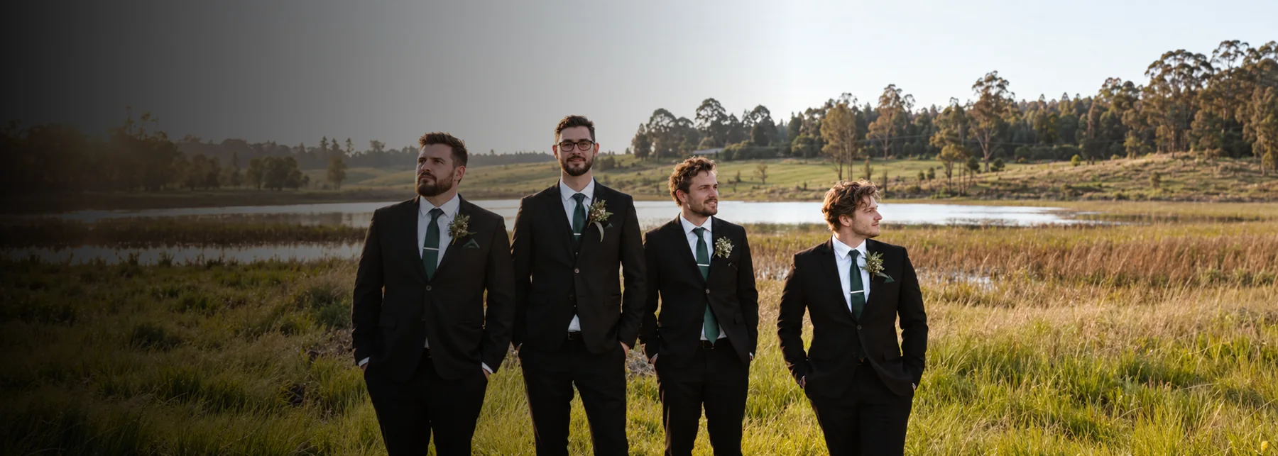 Four groomsmen in black suits with green ties, in a field at sunrise